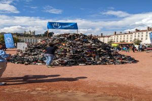 Gruppe von Menschen versammelt um einen Haufen Schuhe auf einem Feld mit Gebäuden, Bäumen und bewölktem Himmel im Hintergrund bei einem internationalen Schuh-Recycling-Event.