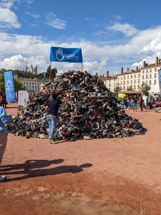 Gruppe von Menschen versammelt um einen Haufen Schuhe auf einem Feld mit Gebäuden, Bäumen und bewölktem Himmel im Hintergrund bei einem internationalen Schuh-Recycling-Event.