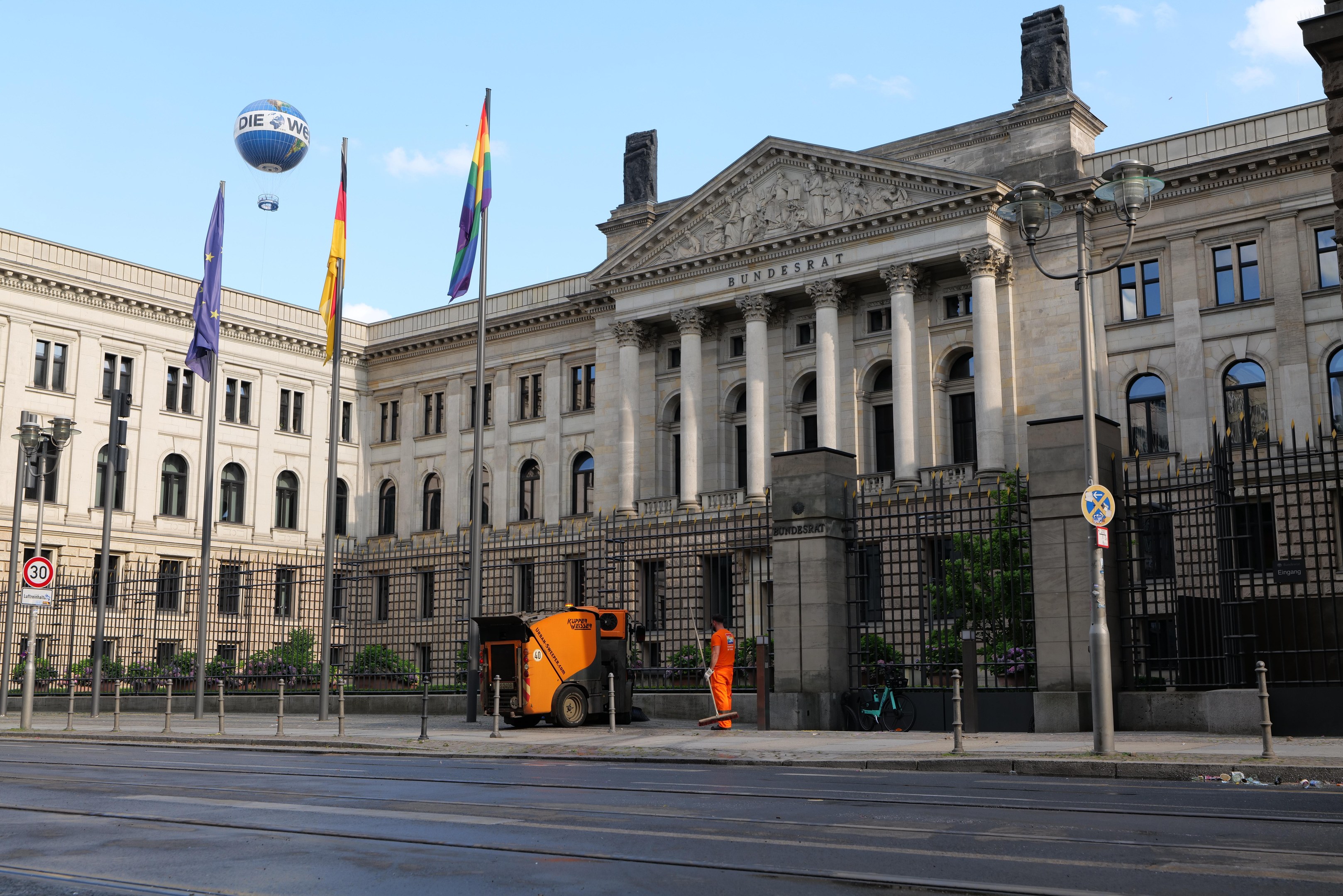 Großes modernes Gebäude mit zahlreichen Fenstern, identifiziert als der Bundestag in Berlin, Deutschland, umgeben von Straßeninfrastruktur, Fußgängern und Flaggen, unter einem bewölkten Himmel.