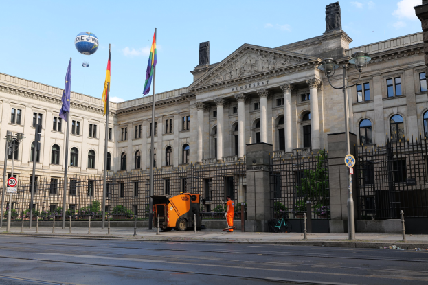Großes modernes Gebäude mit zahlreichen Fenstern, identifiziert als der Bundestag in Berlin, Deutschland, umgeben von Straßeninfrastruktur, Fußgängern und Flaggen, unter einem bewölkten Himmel.
