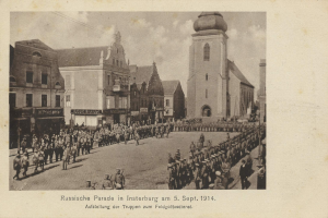 Ein Schwarz-Weiß-Foto eines Umzugs in Insterburg am 5. September 1914 mit vielen Menschen, Gebäuden im Hintergrund, einem bewölkten Himmel und Text unten.