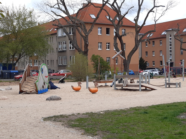 Spielplatz mit einer Rutsche, Schaukeln und anderen Geräten, umgeben von Gras, Bäumen, Gebäuden, Fahrzeugen und einem bewölkten Himmel.