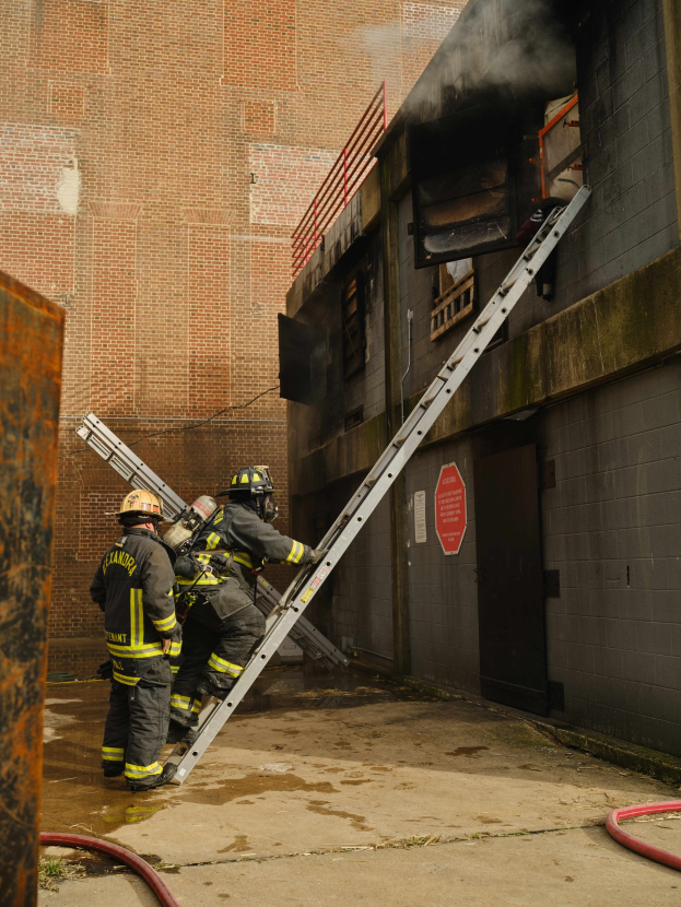 Feuerwehrleute mit Helmen klettern an einer Leiter zu einem rauchgefüllten Gebäude, mit einem Metallobjekt auf der linken Seite und Rohren auf dem Boden.