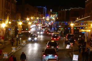 Eine belebte Straße mit verschiedenen Fahrzeugen wie Autos, Lastwagen und Lieferwagen, gesäumt von Gebäuden auf beiden Seiten, mit Fußgängern und Wartenden, Schildern, Werbetafeln, Laternenmasten, Dekorationslichtern und Bäumen.