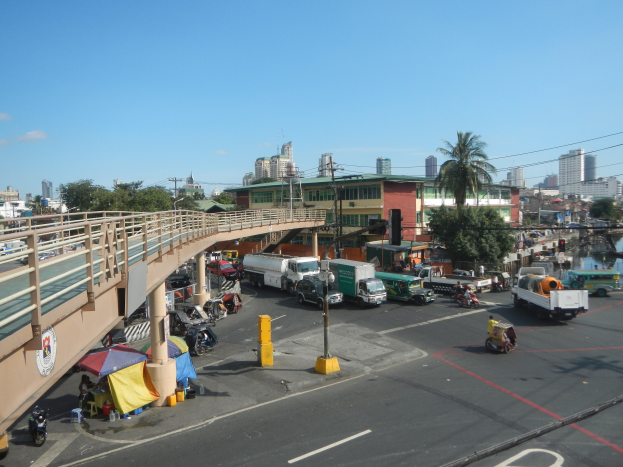 Verkehrsreiche Straße mit Schwerlastverkehr, Föülern, Zelten und Gebäuden in der Nähe einer Brücke über Wasser unter einem bewölkten Himmel.
