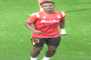 Emmanuel Adebayor, ein Mann in einem roten Hemd und schwarzen Shorts, geht auf einem Fußballfeld mit zwei Personen im Hintergrund.