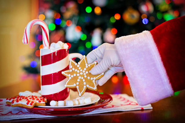 Mann in Santa Claus Mütze mit einer Tasse heißer Schokolade und einem Zuckerstange haltend, mit einem Teller mit Keksen und einem Tuch auf dem Tisch, vor einem Weihnachtsbaum und einer Wand.