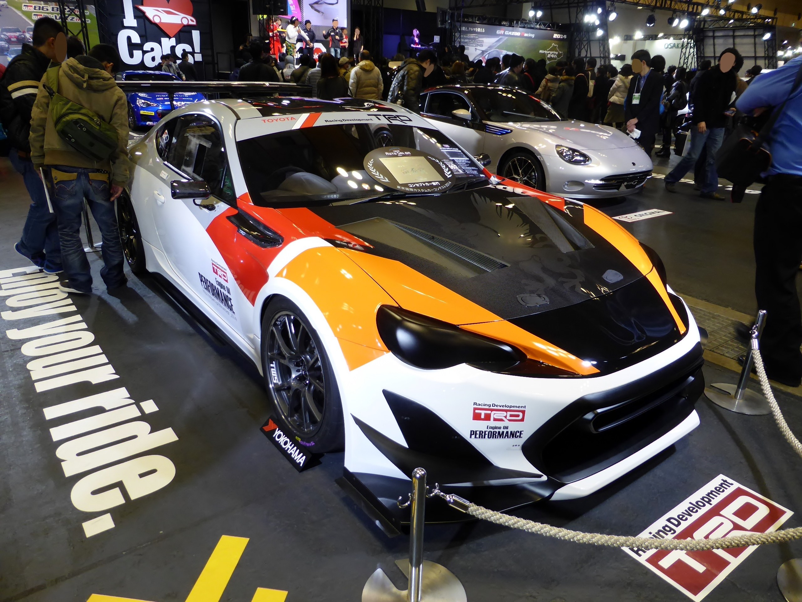 Eine Gruppe von Menschen, die um ein Rennfahrzeug auf der Tokyo Motor Show stehen, umgeben von Absperrpfosten und Seilen, sowie Informationsschildern im Hintergrund unter beleuchteter Decke.