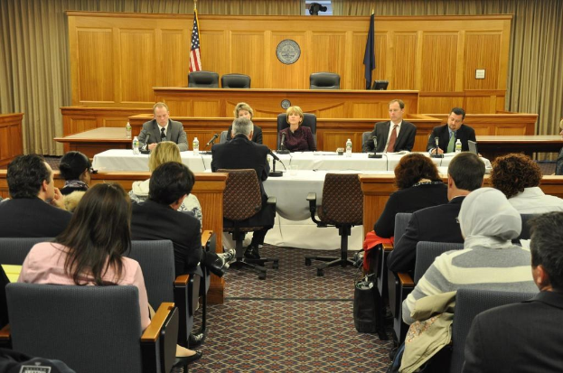 Ein Versammlungsraum mit Menschen auf Stühlen, zwei ausländischen Flaggen, einem orangefarbenen Schreibtisch, Wasserflaschen und einem Mikrofon.