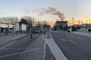 Straßenszene mit Verkehrskegeln, einem Bus, Laternenmasten, Bäumen, Gebäuden, Kränen und Rauch, der aus einem fernen Gebäude aufsteigt, vor einer Stadtkulisse.