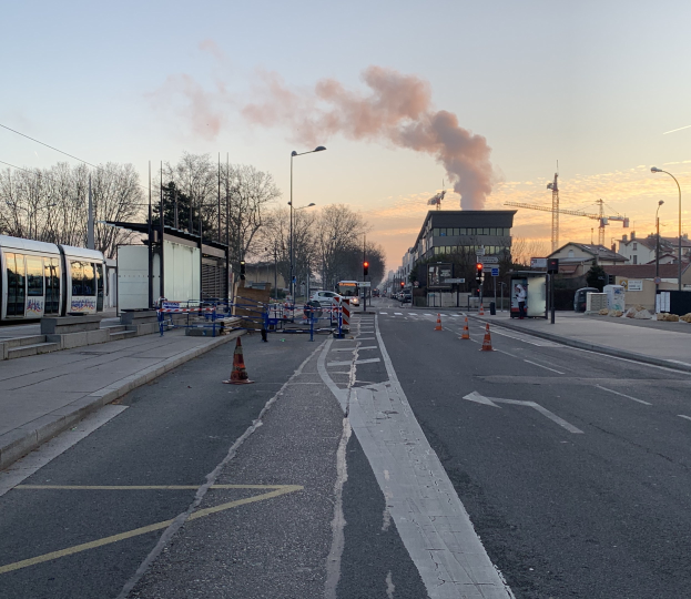 Straßenszene mit Verkehrskegeln, einem Bus, Laternenmasten, Bäumen, Gebäuden, Kränen und Rauch, der aus einem fernen Gebäude aufsteigt, vor einer Stadtkulisse.
