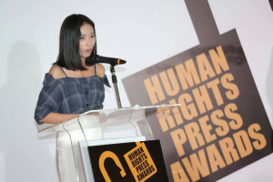 Eine Frau in einem schwarzen Kleid steht an einem Rednerpult mit einem Mikrofon, hält eine Rede oder Präsentation, mit einem Banner dahinter, das "Human Rights Press Awards" lautet.