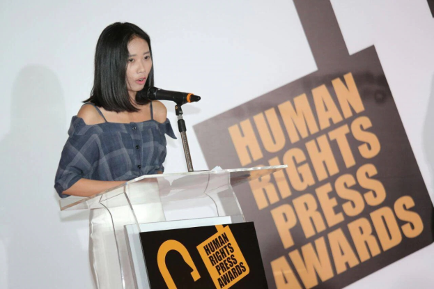 Eine Frau in einem schwarzen Kleid steht an einem Rednerpult mit einem Mikrofon, hält eine Rede oder Präsentation, mit einem Banner dahinter, das "Human Rights Press Awards" lautet.