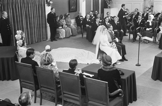 Schwarzes und weißes Foto einer Hochzeitszeremonie mit sitzenden Gästen, einem Mann mit Mikrofon, einer Frau mit einem Strauß, einem Podium mit Mikrofon, Blumenschmuck, einem Vorhang, einem gerahmten Foto an der Wand und einer Tür im Hintergrund.