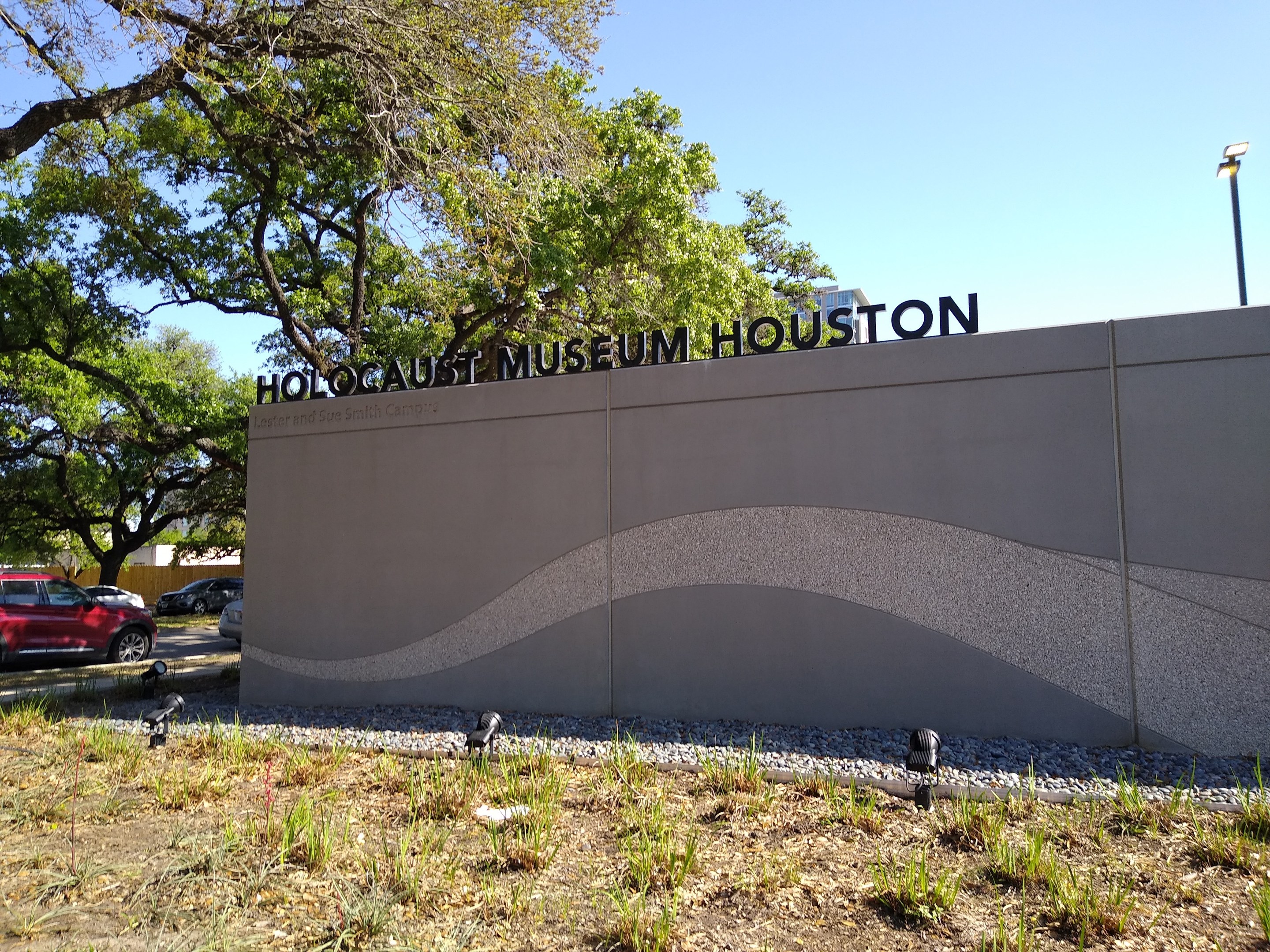 Außenansicht des Houstoner Holocaust Museums mit einer Gedenkwand, Steinen, Vegetation, geparkten Autos, einem Straßenpfahl und einem bewölkten Himmel.