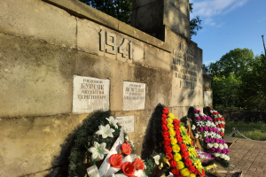 Ein ernster Weltkriegsgedenkmonument mit Blumen darauf, umgeben von einer Wand mit Text und Zahlen, vor einem Hintergrund aus Bäumen und einem bewölkten Himmel.