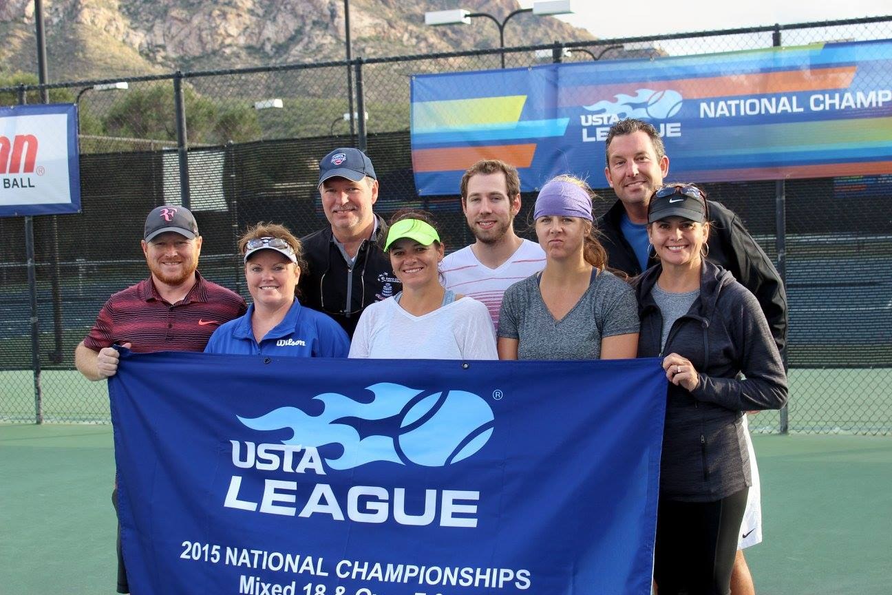 Gruppe von Menschen auf einem Tennisplatz mit einem "2015 USTA National Championships"-Banner, einem Zaun, Bannern, Laternenmasten, Büschen, Bergen und einem klaren blauen Himmel im Hintergrund.