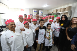 Eine Gruppe von Menschen in einer Küche, die fröhlich posieren und fotografiert werden, einige tragen Mützen, Utensilien links, Regale mit Tellern und Schüsseln im Hintergrund und Deckenleuchten.