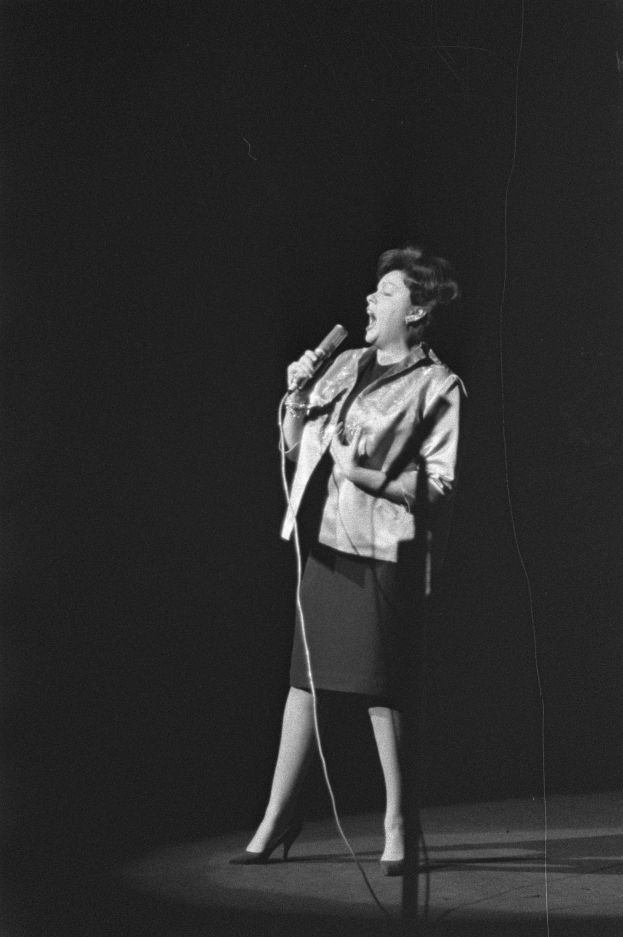 Eine Frau in einem schwarzen und weißen Outfit aus den 1950er Jahren singt fröhlich in ein Mikrofon auf einer von einem Scheinwerfer beleuchteten Bühne.