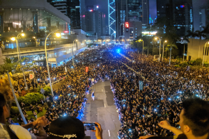 Eine große Menschenmenge steht nachts auf einer Straße, beleuchtet von Straßenlaternen, einige halten dabei Handys hoch während einer Hongkonger Protestaktion.