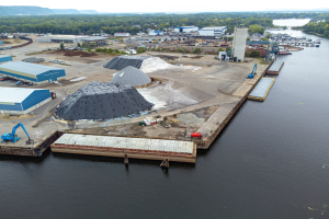 Luftaufnahme eines großen Gewässers mit einem zentralen Kai, umgeben von Gebäuden, Schuppen, Fahrzeugen, Kränen und anderen Strukturen, mit Bäumen, Hügeln und einem klaren blauen Himmel im Hintergrund.