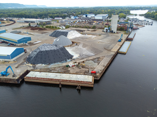 Luftaufnahme eines großen Gewässers mit einem zentralen Kai, umgeben von Gebäuden, Schuppen, Fahrzeugen, Kränen und anderen Strukturen, mit Bäumen, Hügeln und einem klaren blauen Himmel im Hintergrund.