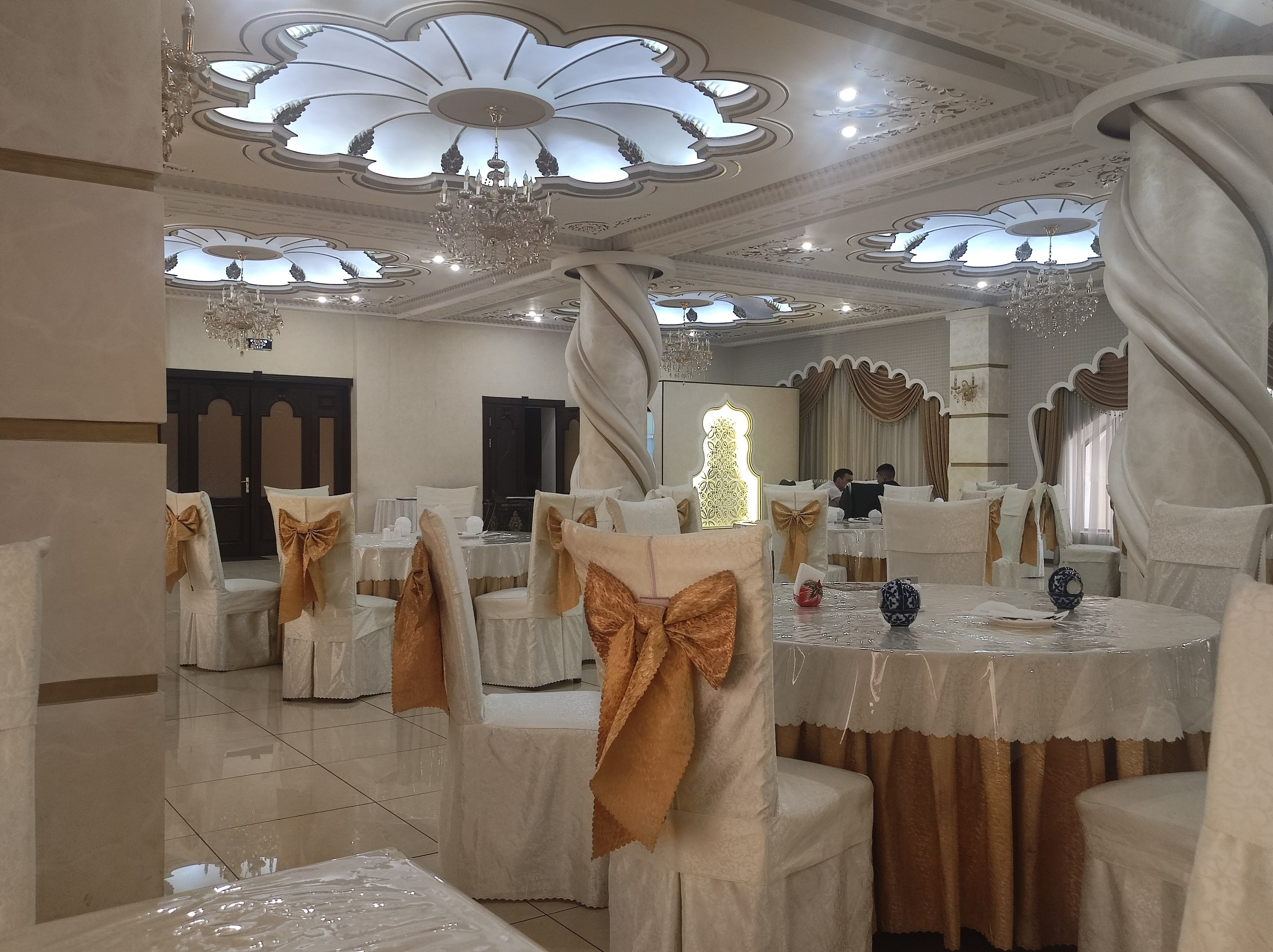 Großer Raum mit Tischen und Stühlen für eine Hochzeitsfeier eingerichtet, Stühle mit Tüchern drapiert und mit dekorativen Accessoires versehen, Wände mit Türen und Vorhängen geschmückt, Decke mit Lichtern und Kronleuchtern dekoriert.