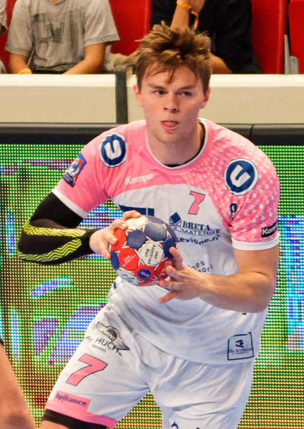 Ein Mann in einem pinken Hemd mit einem Fußball, daneben die Hand einer anderen Person, während im Hintergrund Menschen auf Stühlen sitzen.