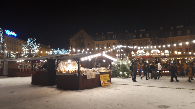 Ein verschneiter Weihnachtsmarkt bei Nacht mit Menschen, Buden und beleuchteten Gebäuden unter einem wolkenverhangenen Himmel.