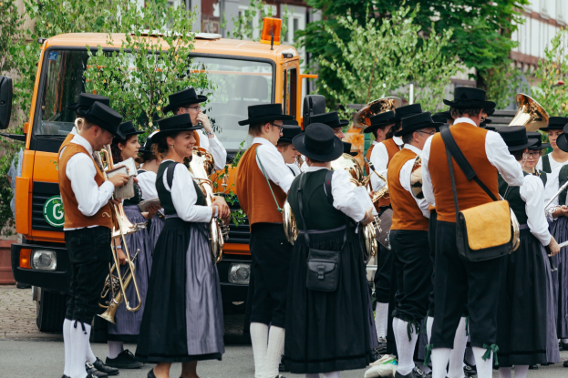 Gruppe von Menschen in traditioneller bayrischer Tracht, die eine Straße entlangmarschieren, Musikinstrumente spielen und Taschen tragen, mit Bäumen, Gebäuden und einem geparkten Fahrzeug im Hintergrund.