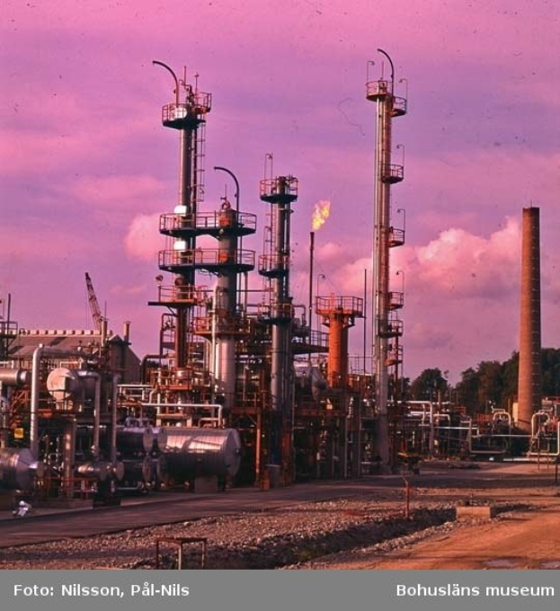 Ölraffinerie mit ausgedehnten Rohrleitungen, Ventilen, Metallstrukturen, Containern, Bäumen, einem Gebäude, einem Kran und einem bewölkten Himmel, mit Text unten.