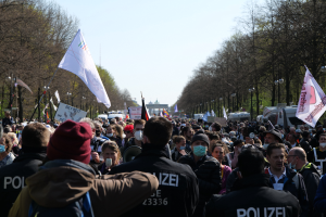 Eine große Gruppe von Menschen steht vor Polizeibeamten, einige tragen Mützen und Masken, während sie auf einer Demonstration in Berlin, Deutschland, mit Schildern, Fahnen, Laternenmasten, Bäumen, Fahrzeugen, einem Gebäude und dem Himmel im Hintergrund demonstrieren.