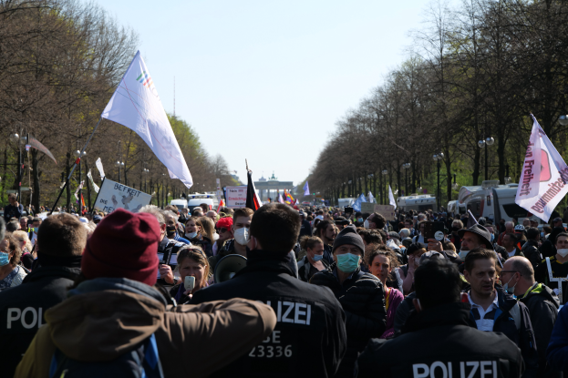 Eine große Gruppe von Menschen steht vor Polizeibeamten, einige tragen Mützen und Masken, während sie auf einer Demonstration in Berlin, Deutschland, mit Schildern, Fahnen, Laternenmasten, Bäumen, Fahrzeugen, einem Gebäude und dem Himmel im Hintergrund demonstrieren.