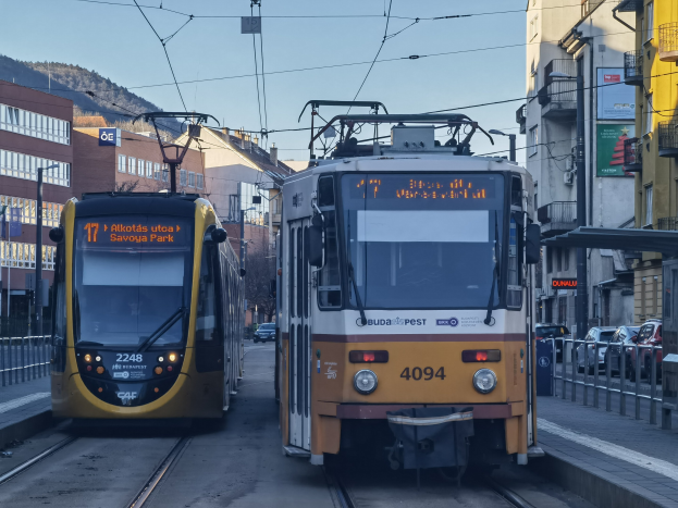 Zwei gelbe und weiße Straßenbahnen fahren auf Stadtgleisen neben hohen Gebäuden, mit Fahrzeugen auf der Straße, Geländern auf beiden Seiten und einem Hintergrund mit Gebäuden, Strommasten, Schildern, Bäumen, einem Hügel und Himmel.
