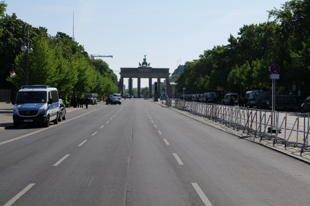Ein Polizeiwagen steht am Straßenrand vor dem Brandenburgertor in Berlin, Deutschland, mit Absperrungen, Schildern, Bäumen und Laternenmasten im Hintergrund und einer bewölkten Himmel.