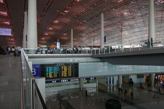 Ein Innenblick in einen Flughafen mit vielen Menschen, die herumlaufen, einige tragen Taschen, Geländer, Säulen, Schilder mit Text und Deckenleuchten.