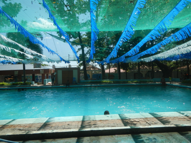 Ein großes Schwimmbad mit blauen und grünen Bändern an der Decke, umgeben von Häusern, Bäumen und einem klaren blauen Himmel, mit Menschen, die das Wasser und die Sonne genießen.