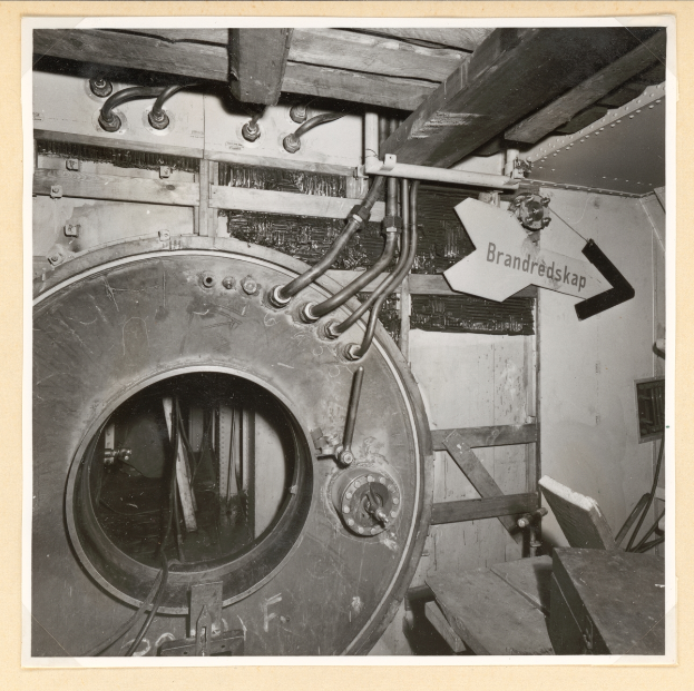 Ein Schwarz-Weiß-Foto einer großen Metallbanktresor mit rundem Fenster, umgeben von Rohren und einer Tafel mit Text, mit verschiedenen Gegenständen auf der rechten Seite verstreut.