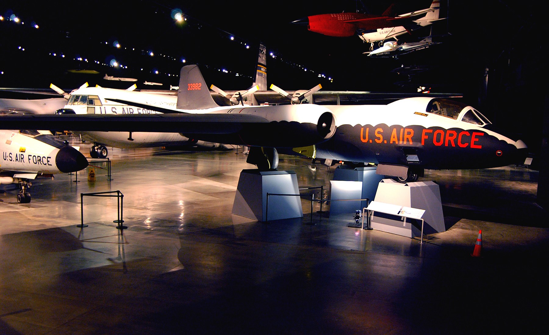 Eine Gruppe von Kampfflugzeugen in einem Museum mit St├Ąnden, Verkehrskegeln und anderen Bodenobjekten ausgestellt, beleuchtet gegen einen dunklen Hintergrund.