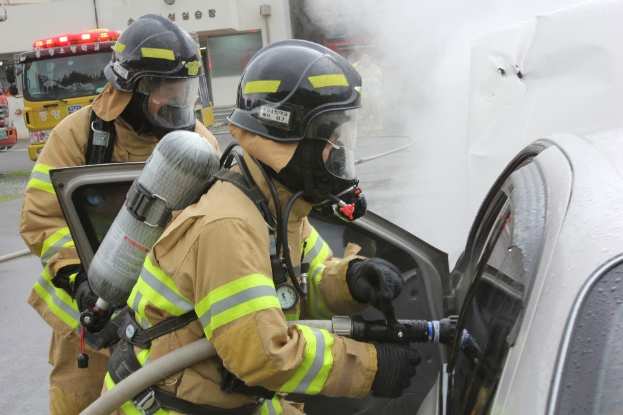 Zwei Feuerwehrleute in Schutzausrüstung verwenden einen Schlauch, um ein brennendes Auto zu löschen, mit Rauch, der daraus aufsteigt, während Fahrzeuge und ein Gebäude im Hintergrund zu sehen sind.