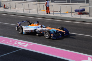 Ein Mann fährt ein Formelauto auf einer Rennstrecke, mit einem Helmträger auf dem Fahrersitz, während eine Person auf dem Gehweg im Hintergrund steht, in der Nähe eines Zauns und eines Feuerlöschers.