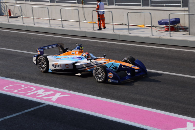 Ein Mann fährt ein Formelauto auf einer Rennstrecke, mit einem Helmträger auf dem Fahrersitz, während eine Person auf dem Gehweg im Hintergrund steht, in der Nähe eines Zauns und eines Feuerlöschers.