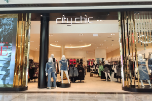 Ein stylischer Bekleidungsladen in einem Einkaufszentrum mit Schaufensterpuppen in der neuesten Mode, Kleidung an Bügeln, einer Säule, einem Schild mit Text und einer Decke mit Lampen sowie einem Fliesenboden.