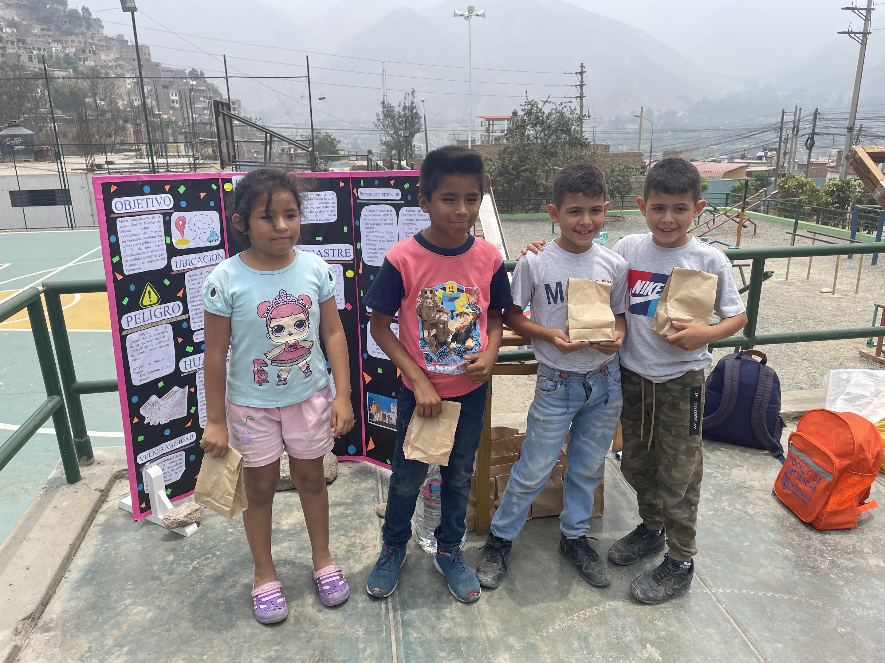 Eine Gruppe von Kindern hält Papiertüten in den Händen und steht auf einem Basketballfeld, daneben liegen Tüten und Gegenstände auf dem Boden, links eine Tafel mit Text und im Hintergrund Zäune, Pfosten und Gebäude.