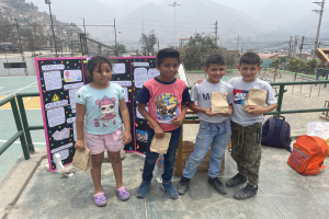Eine Gruppe von Kindern hält Papiertüten in den Händen und steht auf einem Basketballfeld, daneben liegen Tüten und Gegenstände auf dem Boden, links eine Tafel mit Text und im Hintergrund Zäune, Pfosten und Gebäude.
