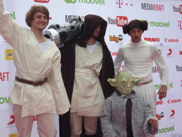 Eine Gruppe von Menschen in Star-Wars-Kostümen, die auf einem roten Teppich posen, wobei einer ein Objekt in der Mitte hält und ein Banner mit Text und Logos im Hintergrund zu sehen ist.