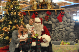 Eine Frau, Kinder und ein Mann in einem Santa-Klaus-Kostüm mit Brille und Handschuhen stehen neben einem geschmückten Weihnachtsbaum, einer Wand mit Steinen, Spielzeugen, Dekorationen, einem Korb mit Gegenständen und Lichtern im Hintergrund. Ein Junge hält ein Päckchen.