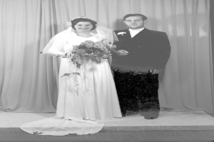 Ein Schwarz-Weiß-Foto eines Brautpaares auf einer Bühne, wobei die Braut einen Strauß hält und der Bräutigam einen Anzug trägt und ein Vorhang dahinter zu sehen ist.