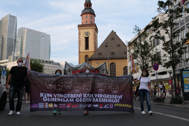 Eine Gruppe von Menschen mit Masken, die eine Straße entlanggehen und ein Transparent halten, mit einem geparkten Auto auf der linken Seite, Gebäuden, Bäumen, Schildern, Masten, einem Uhrenturm und einem klaren blauen Himmel im Hintergrund.