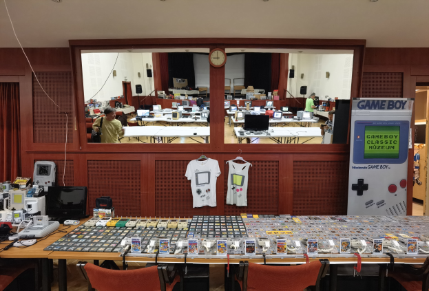 Ein Raum voller Tische und Stühle mit verschiedenen Gegenständen, darunter T-Shirts, Monitore und elektronische Geräte, mit einer Uhr an der Wand und einem Eingang zu einem anderen Raum im Hintergrund, Teil des Nintendo Game Boy Classic Museums.